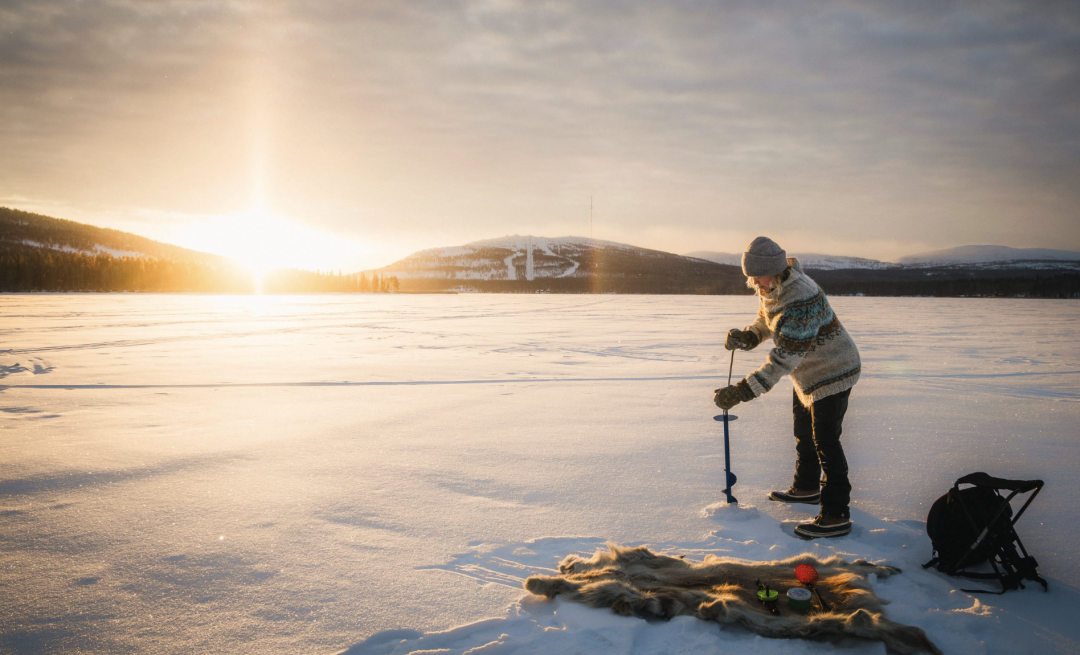 A person drilling a hole to the ice of Pyhäjärvi with a bright sunlight shining through the forest in the back