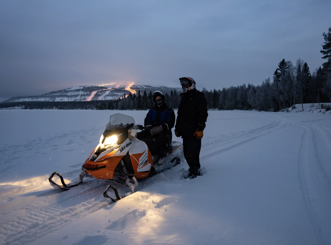 Snowmobile parked on the ice of Pyhäjärvi with a driver and a guide standing next to it, with lights of Pyhä ski slopes in the horizon