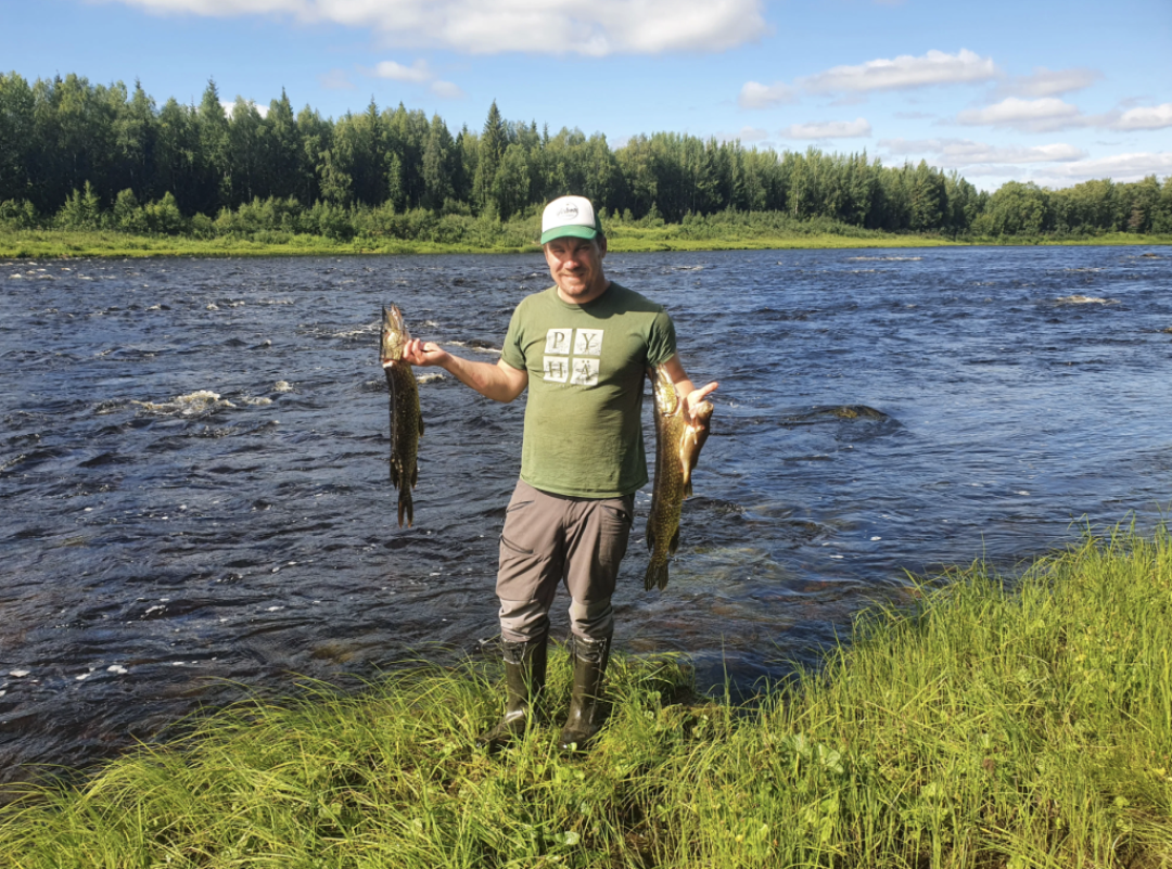 Happy man holding two pikes that he just catched from the raging river behind him with summer greenery under his feet and in the forest behind the river, blue sky above 