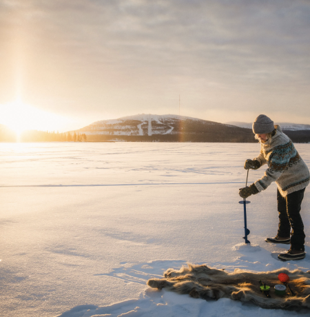 A person drilling a hole to the ice of Pyhäjärvi with a bright sunlight shining through the forest in the back