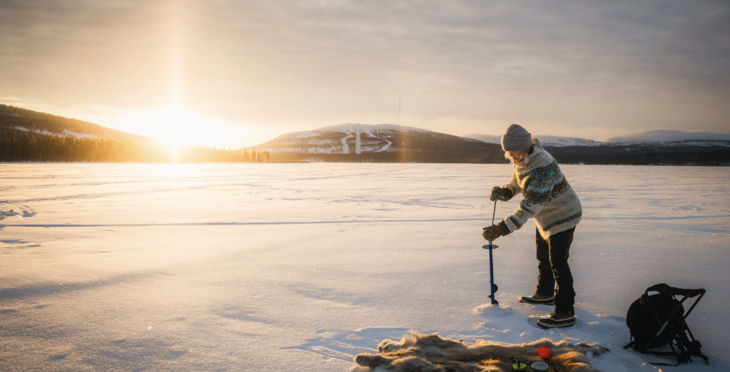 A person drilling a hole to the ice of Pyhäjärvi with a bright sunlight shining through the forest in the back